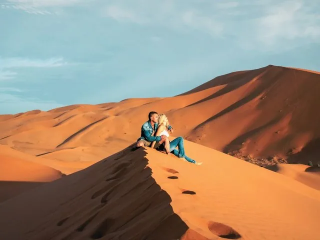 A couple sits together on a high sand dune enjoying the sun