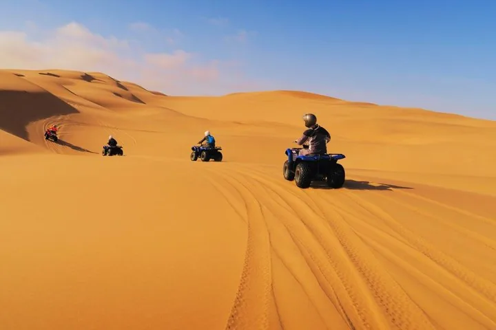 Three people riding quad bikes across the golden sand dunes